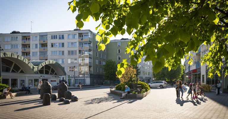 Solig vy över ett torg med några mörkbruna lägre statyer, en grönskande rabatt där någon vilar på en bänk och blad som hänger ner från ett träd i förgrunden, i bakgrunden ett lägre hus med bågformat tak och längst bak högre flerfamiljshus i ljus kulör.