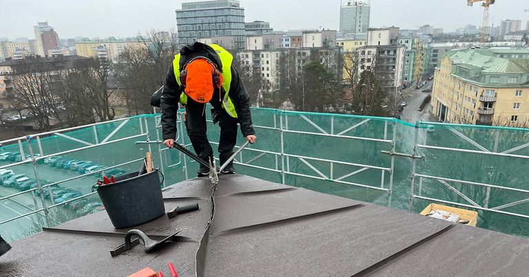 En person i arbetskläder och orange hjälm står lutad neråt och falsar samman plåten på ett plåttak, i bakgrunden syns stadsbilden med flera flerfamiljshus uppifrån.
