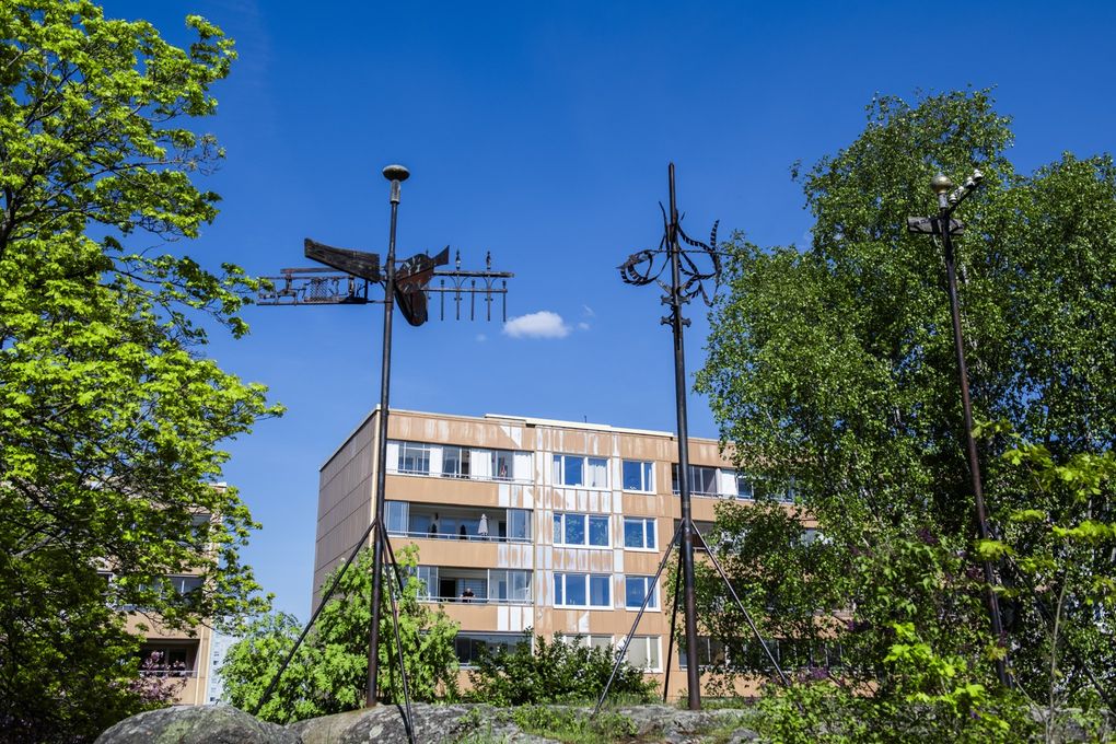 Konstverk ståendes på stenar bland grönska och ett gult lägenhetshus i bakgrunden.